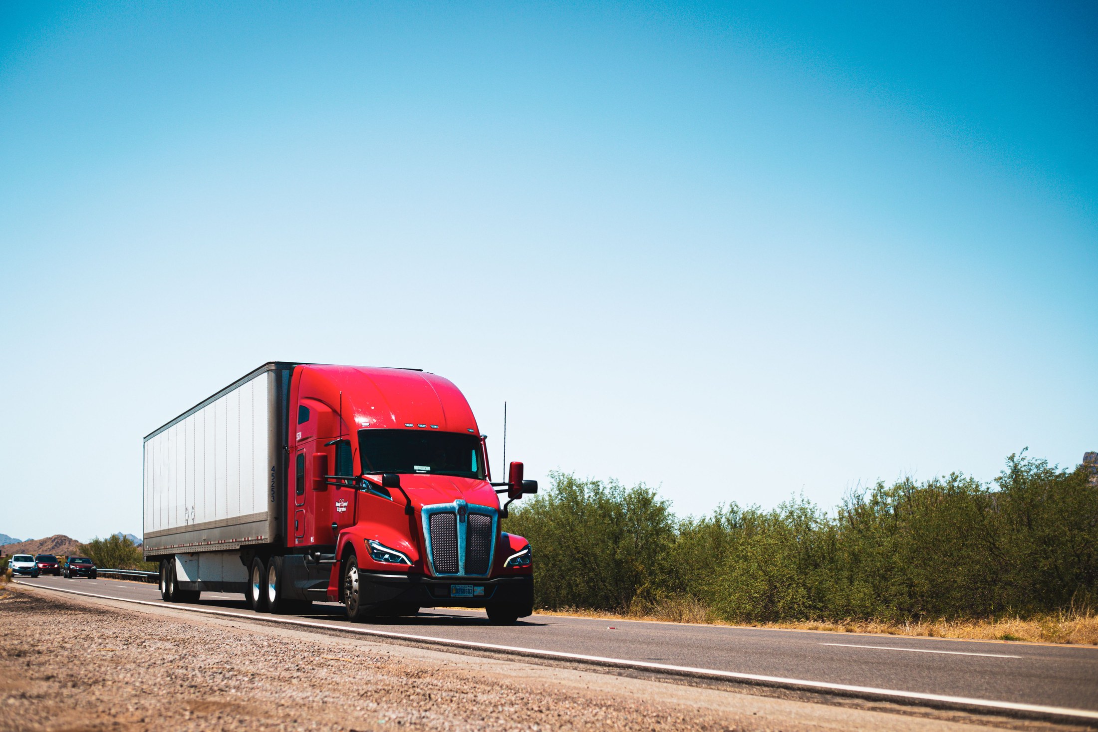 Red conventional-cab semi truck hauling a white dry van trailer on a highway.
